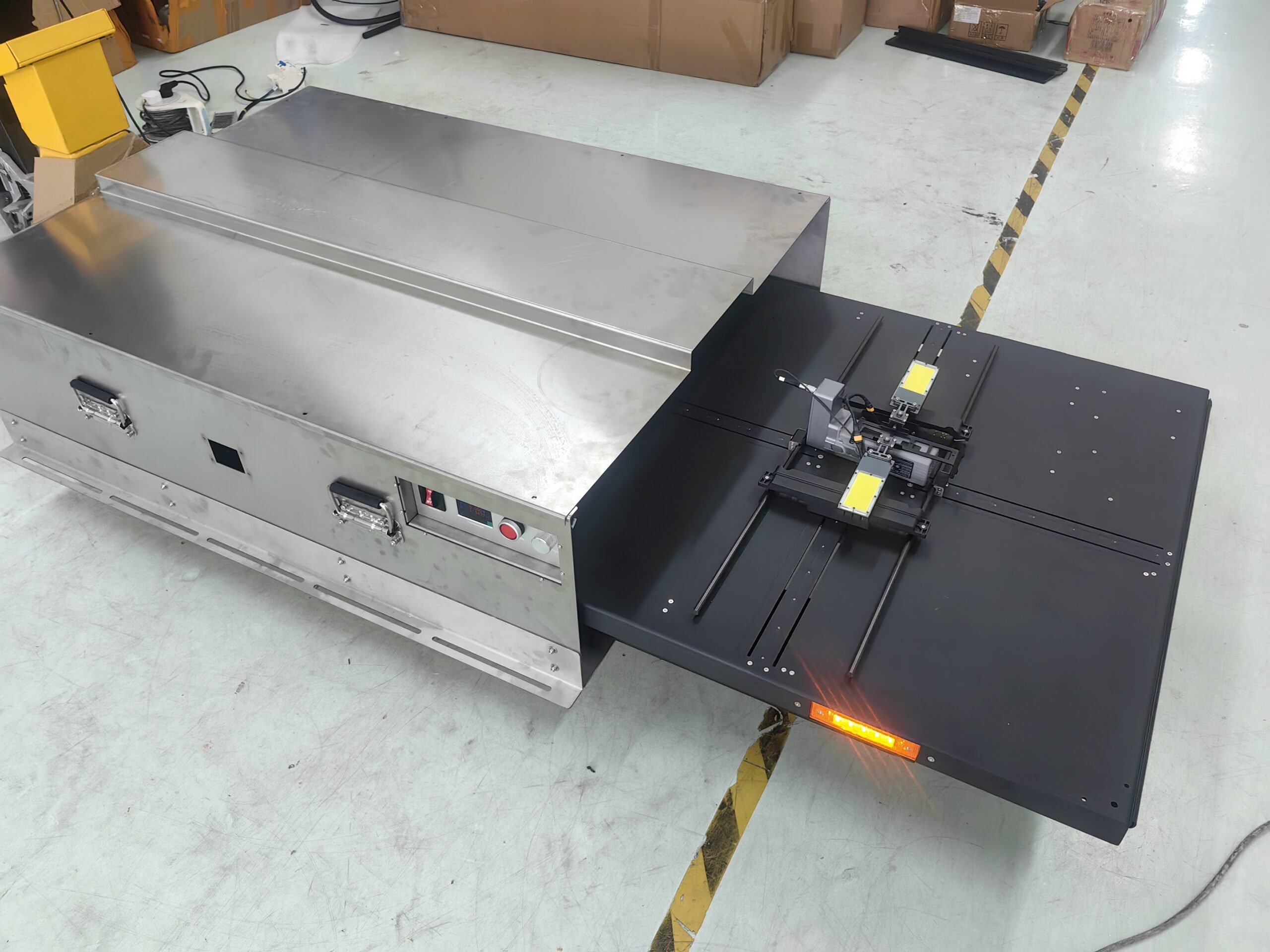 Stainless steel industrial machine in a warehouse, with a black control panel and orange indicator lights, surrounded by cardboard boxes and yellow/black floor tape.