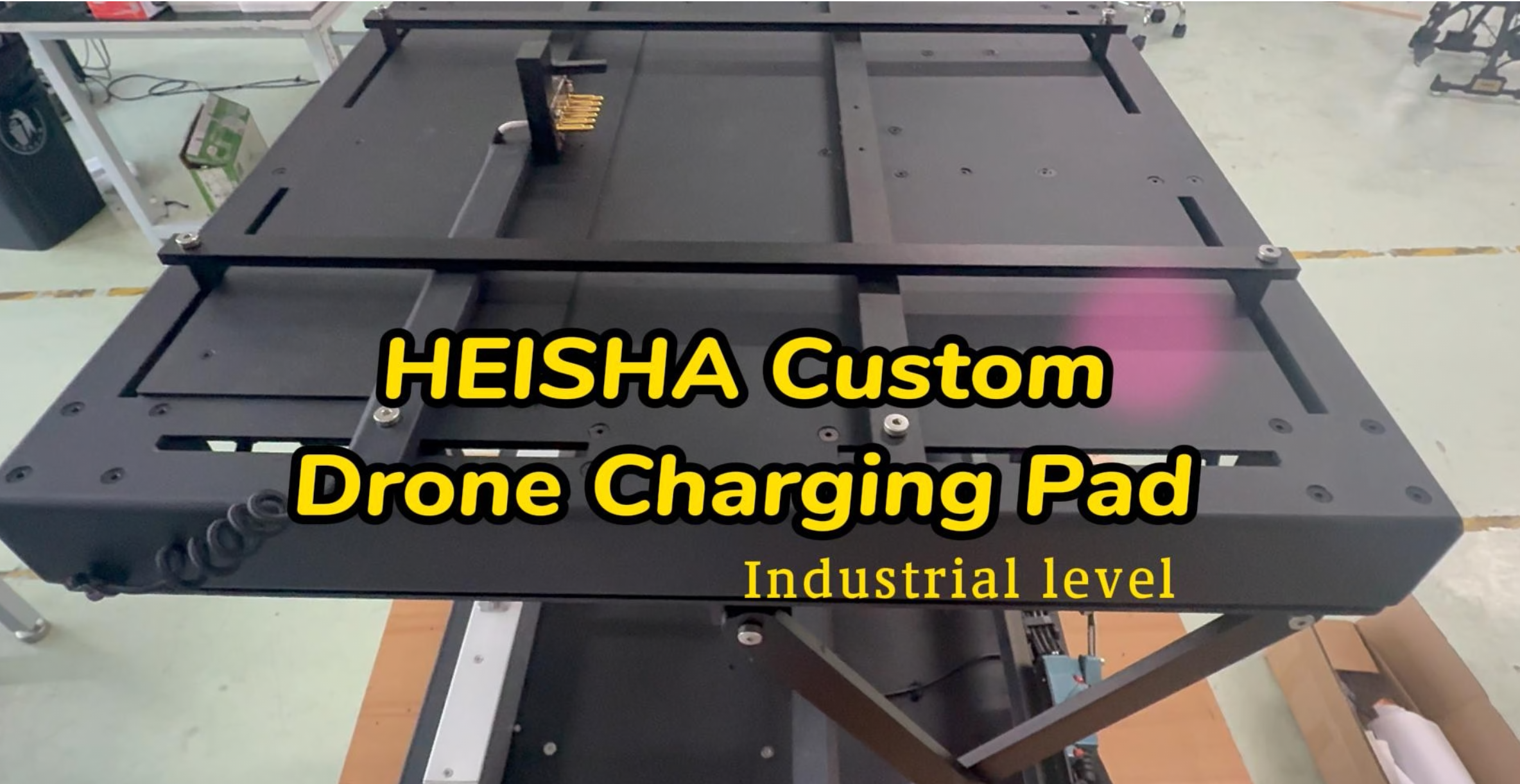 Industrial drone charging pad on a heavy metal frame in a workshop, with bold yellow text reading 'HEISHA Custom Drone Charging Pad'